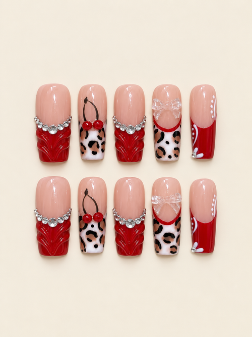 Ten glossy square press-on nails in red and nude with sculpted red accents, raised cherries, clear bows, leopard spots, and rhinestones